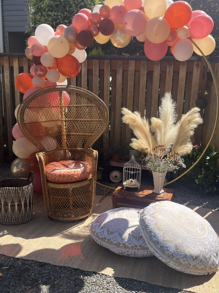 Boho baby shower setup with peacock chair, pampas grass, and balloon arch